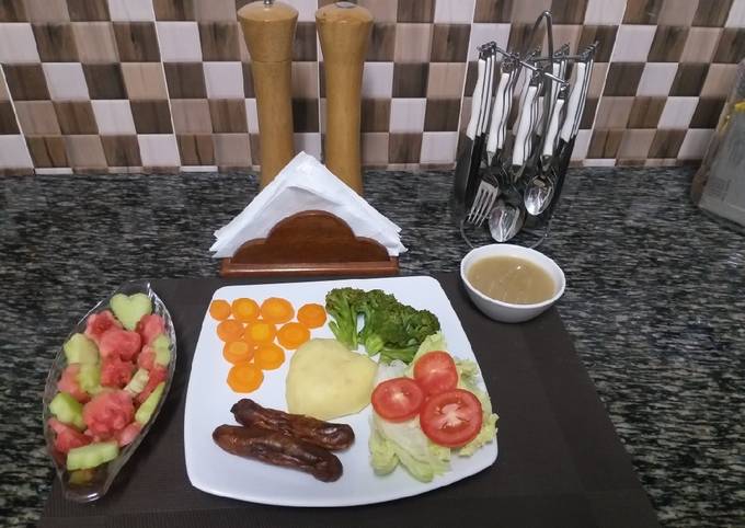 A picture of Mash Potatoes with some steamed veggies,sausages,fruit salad.